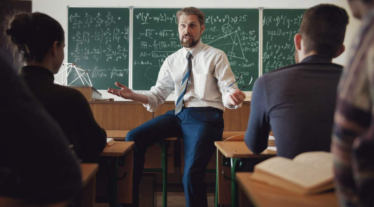 Back view of group of students attending math class and listening to teacher who sits on top of desk